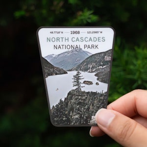 May include: A North Cascades National Park sticker with a scenic view of a lake and mountains. The sticker is gray with white text and coordinates: 48.7718° N - 1968 - 121.2985° W.
