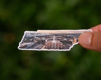 May include: A sticker in the shape of Tennessee, featuring an aerial view of Murfreesboro. The sticker shows a courthouse with a clock tower, surrounded by buildings and parking lots. The word "Murfreesboro" is written in cursive.