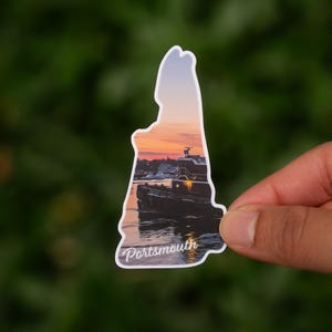 May include: A sticker in the shape of New Hampshire, showing a sunset over a harbour with a boat. The word "Portsmouth" is written in a script font at the bottom. The sticker is held by a hand.
