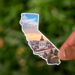 May include: A sticker in the shape of California, featuring a scenic view of Pasadena. The sticker has a white border and the word "Pasadena" in script. The background shows a sunset over buildings and trees.