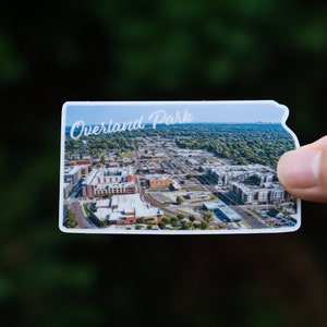 May include: A white sticker with an aerial view of Overland Park, Kansas. The sticker is shaped like the state of Kansas and has the text "Overland Park" in white cursive lettering.