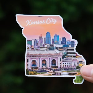 May include: A sticker in the shape of Missouri, featuring the Kansas City skyline and the text "Kansas City". The sticker shows a sunset backdrop and a detailed illustration of the city's architecture.