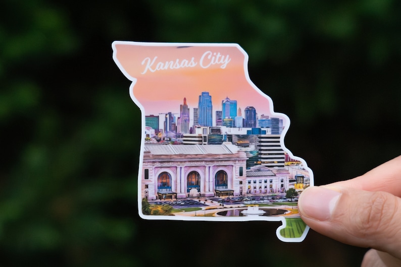 May include: A sticker in the shape of Missouri, featuring the Kansas City skyline and the text "Kansas City". The sticker shows a sunset backdrop and a detailed illustration of the city's architecture.