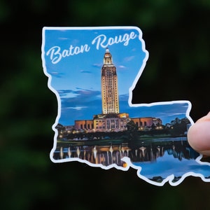 May include: A sticker of the state of Louisiana with a blue sky and a cityscape of Baton Rouge, Louisiana. The state capitol building is the most prominent feature in the image. The text "Baton Rouge" is at the top of the sticker.