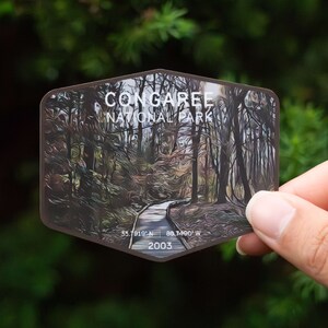 May include: A brown and white sticker with a photo of a path through a forest. The sticker says "Congaree National Park" and includes the coordinates "33.7919° N, 80.7400° W" and the year "2003".