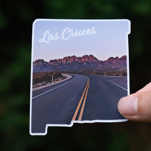 May include: A sticker featuring a scenic view of a winding road leading towards a mountain range at sunset. The sticker is shaped like the state of New Mexico and has the text "Las Cruces" at the top.