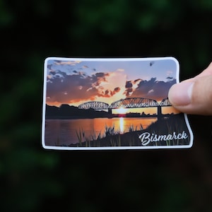 May include: A sticker featuring a sunset over a river with a bridge in the foreground. The text "Bismarck" is printed in white at the bottom of the sticker.