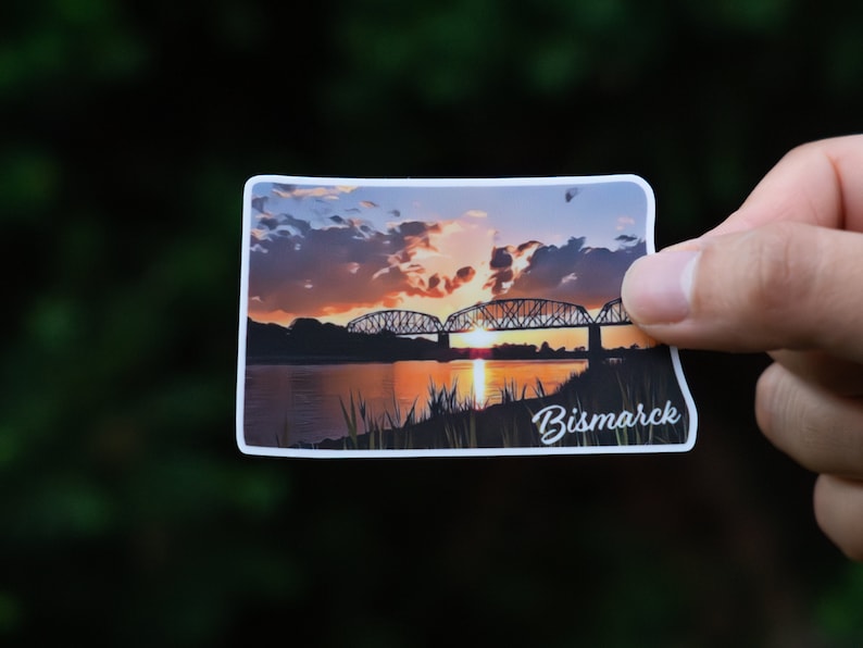 May include: A sticker featuring a sunset over a river with a bridge in the foreground. The text "Bismarck" is printed in white at the bottom of the sticker.