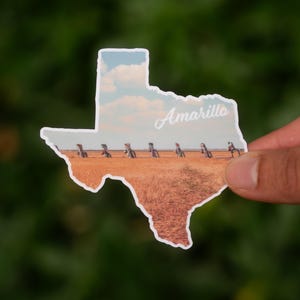 May include: A sticker in the shape of Texas with a landscape scene. The image features the word "Amarillo" in script, a field of wheat, and the Cadillac Ranch art installation. The sky is blue with fluffy clouds.