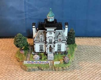 May include: A miniature white and black house with a green roof and a white picket fence. The house is surrounded by green trees and bushes. The house is decorated with red, white, and blue bunting.
