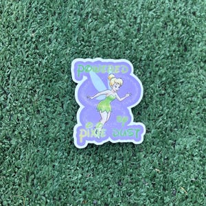 May include: A sticker featuring Tinkerbell in a green dress and wings, with the text "Powered by Pixie Dust." The design has a purple background and white border, set against a green background.