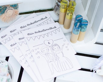 May include: A stack of white coloring books with the text "Mein Hochzeitsmalbuch" and a drawing of a bride and groom. The books are on a white wooden crate, with pencils and other wedding-themed items.