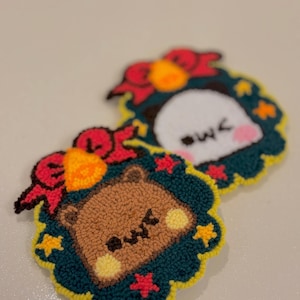 May include: Two handmade, embroidered patches featuring cartoon animals in wreaths. One patch shows a brown bear with a red bow, and the other shows a white penguin with a red bow. The wreaths are green with yellow and red accents.