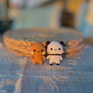 May include: Two handmade crochet bear dolls. One is light brown with yellow cheeks, the other is white with black accents. Both have small arms and legs, and smiling faces. They are standing on a textured white surface.
