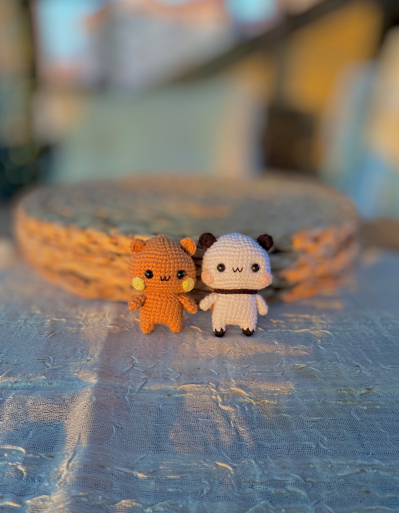 May include: Two handmade crochet bear dolls. One is light brown with yellow cheeks, the other is white with black accents. Both have small arms and legs, and smiling faces. They are standing on a textured white surface.