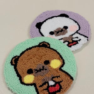 May include: Two round, handmade coasters with cartoon bear designs. One coaster features a brown bear on a light green background, the other a panda bear on a lavender background. Both have black outlines and are made of textured yarn.