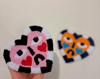 Peut inclure: Deux œuvres d'art textiles en forme de cœur. L'une est rose avec des bordures à carreaux noirs et blancs, des traits noirs et des cœurs rouges. L'autre est orange avec des accents bleus et une bordure à carreaux bleu marine.