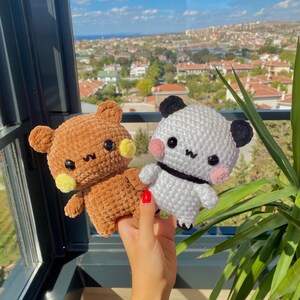 May include: Two crocheted stuffed animals: a brown bear and a panda. The bear has yellow accents and the panda has black ears and a black band. Both have black eyes and are held up in front of a window with a city view.