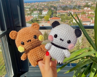 Puede incluir: Dos animales de peluche de ganchillo: un oso marrón y un panda. El oso tiene detalles amarillos y el panda tiene orejas negras y una banda negra. Ambos tienen ojos negros y se sostienen frente a una ventana con vista a la ciudad.