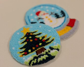 Peut inclure: Deux dessous de verre ronds, bleus clairs, avec des motifs de Noël. L'un représente un sapin de Noël avec une étoile jaune, l'autre un bonhomme de neige. Les dessous de verre sont en matière texturée.