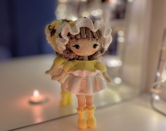 Può includere: Una bambola all'uncinetto con un vestito bianco e giallo e un cappello bianco all'uncinetto. La bambola è in piedi su una superficie bianca con due candele accese.
