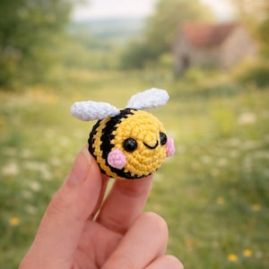 May include: A small, crocheted bee with a yellow body, black stripes, and white wings. The bee has a smiling face with black eyes and pink cheeks. The handmade item is held in front of a blurred green background.