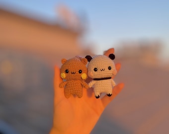 May include: Two small, handmade crochet dolls, one tan and one white, are held in a hand against a soft-focus sunset backdrop. The dolls have black eyes, small noses, and are holding hands. The tan doll has a bear-like appearance, while the white doll resembles a panda.