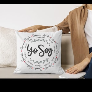 May include: White square pillow with the text "Yo Soy" in the center, surrounded by a wreath and additional text. The pillow is on a white surface, with a person's arm and a book visible in the background.