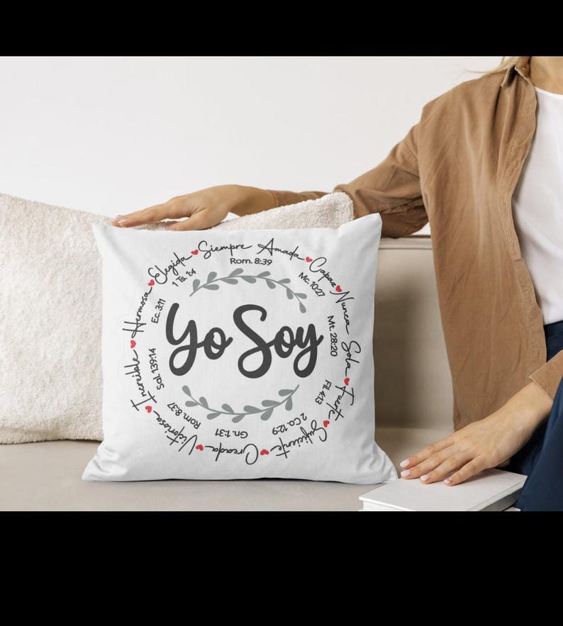 May include: White square pillow with the text "Yo Soy" in the center, surrounded by a wreath and additional text. The pillow is on a white surface, with a person's arm and a book visible in the background.