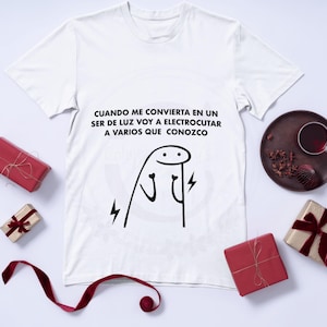 Puede incluir: Camiseta blanca con texto negro que dice "CUANDO ME CONVIERTA EN UN SER DE LUZ VOY A ELECTROCUTAR A VARIOS QUE CONOZCO". El texto está acompañado por un simple dibujo de línea negra de un palo con rayos que salen de sus manos.