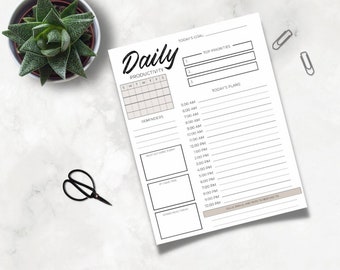 May include: A white daily planner with the word "Daily" in a stylish font. The planner includes sections for top priorities, daily plans, and reminders. A small succulent in a pot, scissors, and paper clips are also in the image.