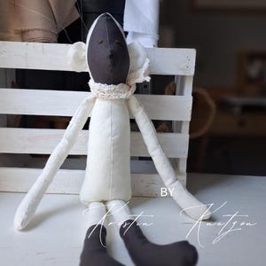 May include: A handmade fabric doll featuring a dark brown head and feet, a cream-colored body, and long arms. The doll wears a small, textured cream-colored crocheted scarf around its neck. The text "BY Kristin Knutzen" is visible on the lower right.