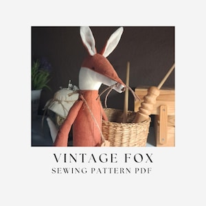 May include: A handmade fabric fox doll with a rust-coloured body, white face, and long ears. The fox has thin brown string whiskers and is positioned near a woven basket and wooden tools. The text VINTAGE FOX SEWING PATTERN PDF is displayed below.