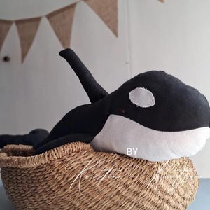 May include: A plush orca toy featuring a black body with a white underbelly and a white eye patch. The stuffed animal has a prominent dorsal fin on its back and is positioned inside a woven, light brown, round basket with a textured surface.
