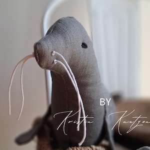 May include: A grey fabric seal plush toy featuring a textured linen-like surface, a small black embroidered eye, and long white string whiskers. The text BY Kristin Knutzen is printed in white on the right side of the seal's body.