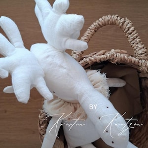 May include: A white fabric stuffed animal with long ears and large hands, featuring a beige ruffled collar around the neck. The toy is positioned inside a woven wicker basket. The text BY KRISTIN KNUTZEN is visible on the toy's surface.
