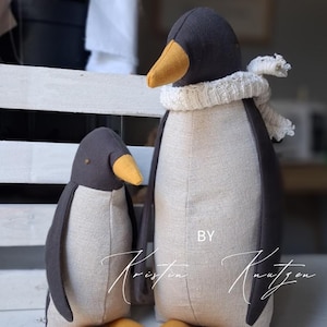 May include: Two stuffed penguin toys made of dark gray and beige fabric with yellow beaks. The larger penguin wears a white knitted scarf around its neck. The text BY Kristin Knutzen is visible in white script across the lower portion of the image.