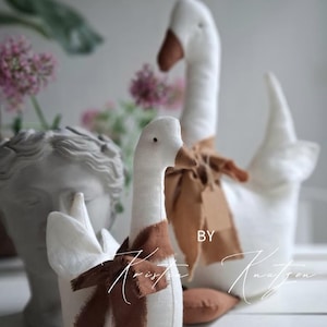 May include: Two white fabric goose figurines with brown beaks and brown fabric ribbons tied around their necks. The geese are positioned in front of a gray bust sculpture. The text "BY KRISTA KNU TZEN" is visible across the lower portion of the image.