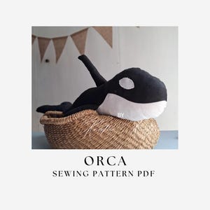 Puede incluir: Un peluche de orca de tela en blanco y negro que descansa dentro de una cesta de paja tejida. El texto ORCA SEWING PATTERN PDF está impreso debajo de la imagen. La orca presenta una aleta dorsal prominente y un parche ocular blanco.