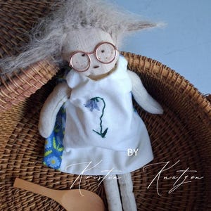 May include: A handmade fabric doll with messy light gray hair and round copper-colored wire glasses. The doll wears a white sleeveless dress featuring a small embroidered blue flower on the front and a blue floral patterned side panel. Text reads BY Kristin Knutzen.