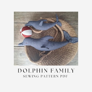 May include: Two grey fabric dolphin plush toys and a small red and white striped ball. The text DOLPHIN FAMILY SEWING PATTERN PDF is printed below the items. The dolphins feature embroidered details on their tails and fins.