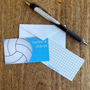 May include: A black and silver pen rests on a wooden surface next to a white envelope and two cards. One card has a volleyball graphic and the text "You're a champ!" The other card has a gray grid pattern. The website www.craftyvd.com is visible.
