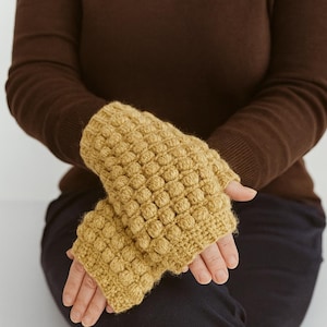 Könnte beinhalten: Senfgelbe Fingerhandschuhe mit einem strukturierten, erhabenen Muster. Die Handschuhe sind aus einem weichen, gestrickten Material gefertigt und bedecken die Hand bis zu den Knöcheln, wobei die Finger frei bleiben. Die Handschuhe werden vor einem braunen Hintergrund gezeigt.