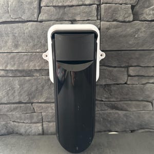 Könnte beinhalten: Ein schwarz-weißer Sicherheitssensor an einer grauen Steinmauer. Der Sensor hat einen rechteckigen schwarzen Körper mit gewölbter Oberseite und ist in einem weißen Rahmen eingefasst. Die Wand hat ein strukturiertes, geschichtetes Aussehen.