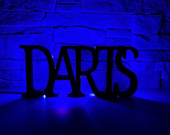 Puede incluir: Letras de madera negras deletreando la palabra "DARTS" sobre un fondo de pared de ladrillo azul.