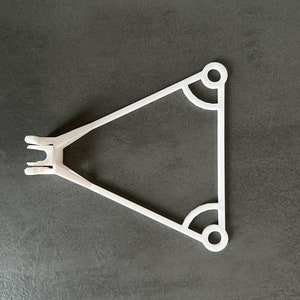 Pode incluir: Cabide de plástico branco com forma triangular e dois furos redondos na parte superior. O cabide tem um gancho de um lado e uma pequena ponta pontiaguda do outro.
