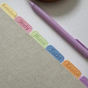Könnte beinhalten: Ein beigefarbenes, stoffbezogenes Notizbuch mit einer Reihe bunter Kunststoff-Registerkarten mit der Beschriftung Revision, Study, Notes, Tasks, Exams und Deadlines sowie einem glatten lavendelfarbenen Stift, der diagonal auf der Seite liegt.