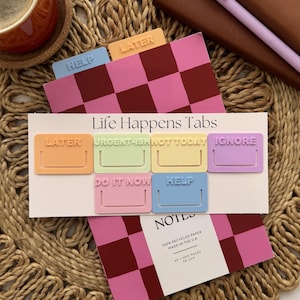 Op de afbeelding: Een set Life Happens-tabbladen in pastelkleuren zoals oranje, groen, geel, paars, roze en blauw. Elk tabblad bevat tekst in reliëf zoals LATER, URGENT-ISH, NOT TODAY, IGNORE, DO IT NOW en HELP op een witte achtergrondkaart.