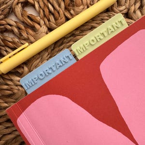 May include: A red, pink and yellow notebook with three blue, light green and yellow bookmarks that say "IMPORTANT" on them. The notebook is on a woven straw surface.