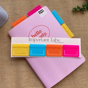 May include: Four colourful plastic page markers with the word "IMPORTANT" embossed on each. The markers are yellow, blue, orange, and pink. The markers are on a pink notebook with the text "hello lovely. Important Tabs by Lovewillowk" printed on the cover.